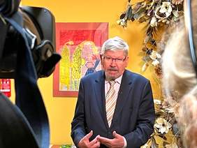 Pokaż zdjęcie: Przed obiektywem kamery stoi mężczyzna. Jest ubrany w elegancki garnitur. Mówi i gestykuluje rękoma. 