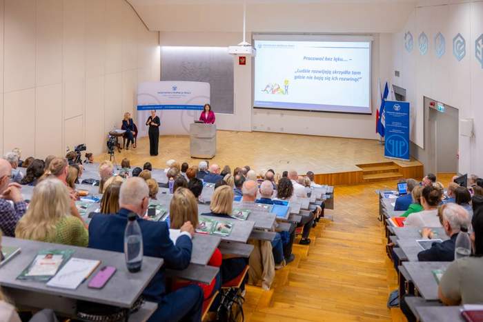 Uczestnicy spotkania podczas wykłądu prelegentki.W tle widać mównicę, prelegentkę, tłumacza języka migowego oraz ekran z wyświetloną prezentacją.