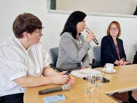 Pokaż zdjęcie: Trzy kobiety siedzą przy stole, jedna przemawia do mikrofonu