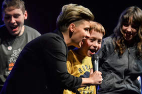 Pokaż zdjęcie: Na zdjęciu 4 śpiewające osoby lekko pochylone do mikrofonu.