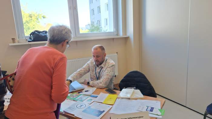 Przy stole siedzi meżczyzna na wózku inwalidzkim - Przedstawiciel Kujawsko-Pomorskiego Oddziału PFRON i rozmawia z kobietą, która przed nim stoi. Przed nim na stole leżą ulotki i broszury PFRON.