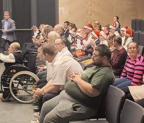 Pokaż zdjęcie: Osoby siedzące na widowni w tym osoby  z niepełnosprawnością, w tle dzieci i młodzież z czerwonymi czapkami świętego Mikołaja