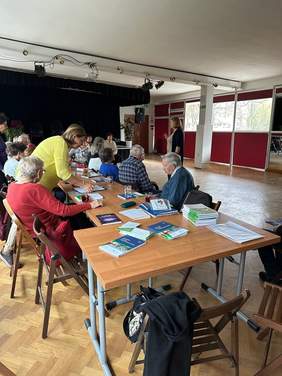 Pokaż zdjęcie: Na stole, wokół którego siedzą ludzie, leżą różne informatory PFRON. W tle widać kobietę prowadzącą spotkanie. 