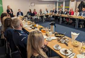 Grupa osób, kobiety i mężczyźni, siedzą przy stole, jedna kobieta stoi, widać, że mówi