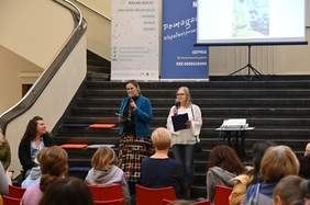 Pokaż zdjęcie: W holu na dużych schodach wejściowych rozchodzących się na boki stoją dwie kobiety. Za nimi na półpiętrze stoją dwa banery i ekran do rzutnika na którym leci prezentacja. Kobiety trzymają w rękach mikrofony i mówią do ludzi siedzących na kr