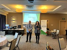 Pokaż zdjęcie: Sala konferencyjna. Po lewej stronie stolik z urządzeniami elektronicznymi na środku wisi ekran do rzutnika z prezentacją z logo PFRON. Obok baner z logo PFRON z napisem wszyscy jesteśmy tacy sami. Na środku stoi mężczyzna i kobieta. On w jasnej ma