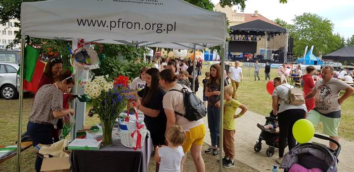 Pokaż zdjęcie: Dwie kobiety stoją pod namiotem z logo PFRON. Na stole przykrytym szarym płótnem rozłożone zostały ulotki oraz materiały informacyjne. Dodatkowo na stole ustawione zostały cztery wazony z bukietami polnych kwiatów oraz kosze wiklinowe z ulotkami i gadżetami z logo PFRON. Wokół stoiska ustawiły się dzieci, które biorą udział w konkursach zorganizowanych przez Oddział Wielkopolski PFRON z zagadnień poruszających tematykę niepełnosprawności. W tle widoczna jest duża scena oraz osoby przemieszczające się pomiędzy stoiskami.