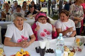 Pokaż zdjęcie: Grupa kilkunastu uśmiechniętych osób siedzi przy stołach, które zostały ustawione w czterech rzędach. Na stolikach znajdują się zarówno kawałki różnych ciast, owoce, jak i napoje.