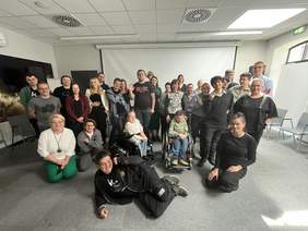 Pokaż zdjęcie: Duża grupa ludzi, w tym osoby na wózkach inwalidzkich i jedna osoba leżąca na podłodze, pozuje do zdjęcia grupowego w jasnym pomieszczeniu.