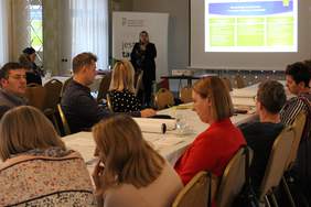 Pokaż zdjęcie: mężczyźni i kobiety patrząna kobietę z mikrofonem w tle prezentacja projektu