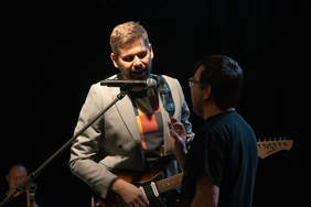 Pokaż zdjęcie: Zdjęcie dwóch artystów na scenie. Mężczyźni stoją do siebie przodem i śpiewają, jeden z nich gra na gitarze.