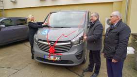 Pokaż zdjęcie: Szary bus, kobieta przecina czerwoną wstążkę. Obok stoi dwóch mężczyzn.