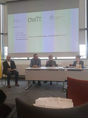 Pokaż zdjęcie: Grupa 4 osób siedzących za stołem prezydialnym, na ekranie wyświetla się prezentacja