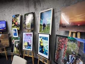 Pokaż zdjęcie: Zdjęcie prezentuje wystawę fotografii. Kilka dużych, oprawionych zdjęć, a także mniejsze odbitki, stoi na sztalugach lub jest opartych o ścianę o surowej, betonowej fakturze. Tematyka zdjęć jest różnorodna, w tym ujęcia natury, portrety i sceny plenerowe.