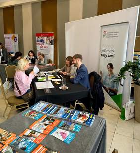 Pokaż zdjęcie: Stoisko Państwowego Funduszu Rehabilitacji Osób Niepełnosprawnych (PFRON) na targach pracy. Przy stole siedzi trójka osób: dwie przedstawicielki PFRON i jeden mężczyzna w garniturze, prowadzą rozmowę z kobietą w różowym swetrze. Na stole leżą ulotki i broszury informacyjne, a w tle widoczne są banery PFRON oraz Poczty Polskiej