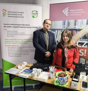 Pokaż zdjęcie: Przy stoliku na którym leżą materiały informacyjno-promocyjne stoją dwie osoby kobieta i mężczyzna. W tle rollup z logo PFRON oaz CIDON.