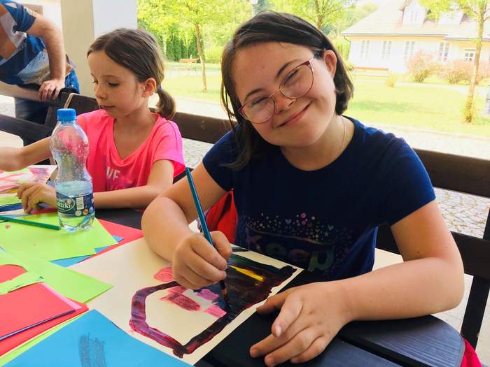 Pokaż zdjęcie: Uśmiechnięta dziewczynka - podopieczna Stowarzyszenia "Tęcza" w takcie warsztatów plastycznych. Siedzi przy drewnianej ławie, maluje farbami.