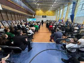 Pokaż zdjęcie: W szkolnej sali gimnastycznej odbywa sie uroczystość. Tłum gości siedzi na krzesłach tyłem do zdjęcia. W tle widać prowadzących uroczystość.  