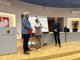 Pokaż zdjęcie: Kobieta w jasnej bluzce i szarej marynarce trzyma mikrofon po obu jej stronach stoją mężczyźni w marynarkach. Za nimi wiszą duże kartki z pismem odręcznym. Po lewej mównica i godło polski. Po prawej w tle herb województwa pomorskiego.