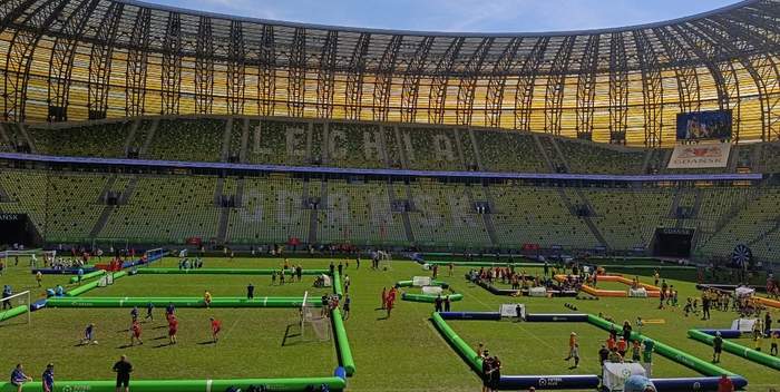Pokaż zdjęcie: Widok wnętrza stadionu Polsat Plus Arena Gdańsk podczas turnieju piłkarskiego. Na murawie rozmieszczono kilka małych boisk z zielonymi bandami, na których grają dzieci i młodzież. Trybuny są częściowo puste, a przez konstrukcję dachu wpada światło słońce.