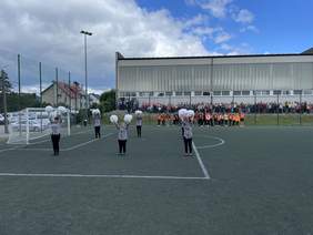 Pokaż zdjęcie: Na sztucznej murawie boiska piłkarskiego przy bramce stoi 6 dziewczyn ubranych w czarne spodnie koszule w biało czarną kratę trzymają w dłoniach białe pompony, za nimi w tle stoją zawodnicy.