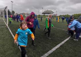 Pokaż zdjęcie: Dzieci i młodzież biegną wokół boiska. Pada deszcz. Zawodnicy mają na sobie peleryny, niektórzy biegną z parasolami.