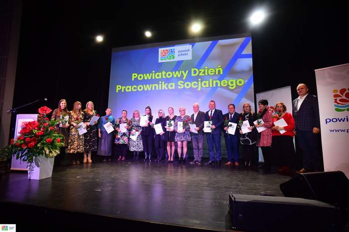 Pokaż zdjęcie: Grupa kilkunastu osób stoi na scenie. Każda a nich trzyma w dłoniach czerwoną różę oraz torbę z logo powiatu pilskiego. Po lewej stronie widoczna jest mównica oraz duży bukiet kwiatów w kolorze czerwonym. W tle możemy dostrzec duży ekran, na którym wyświetlony jest logotyp powiatu pilskiego oraz sformułowanie: „Powiatowy Dzień Pracownika Socjalnego”.