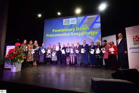Pokaż zdjęcie: Grupa kilkunastu osób stoi na scenie. Każda a nich trzyma w dłoniach czerwoną różę oraz torbę z logo powiatu pilskiego. Po lewej stronie widoczna jest mównica oraz duży bukiet kwiatów w kolorze czerwonym. W tle możemy dostrzec duży ekran, na którym wyświetlony jest logotyp powiatu pilskiego oraz sformułowanie: „Powiatowy Dzień Pracownika Socjalnego”.