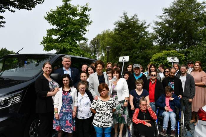 Pokaż zdjęcie: Grupa około trzydziestu osób stoi obok czarnego mikrobusa. Z przodu znajdują się dwie osoby poruszające się na wózku. W tle drzewa liściaste oraz dwa słupki z oznaczeniem miejsca parkingowego dla osób z niepełnosprawnością.