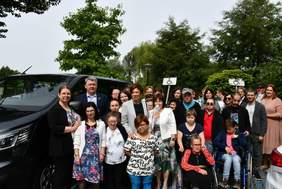 Pokaż zdjęcie: Grupa około trzydziestu osób stoi obok czarnego mikrobusa. Z przodu znajdują się dwie osoby poruszające się na wózku. W tle drzewa liściaste oraz dwa słupki z oznaczeniem miejsca parkingowego dla osób z niepełnosprawnością.