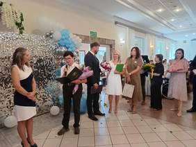 Pokaż zdjęcie: Grupa kilku osób stoi w kolejce do składania życzeń oraz wręczania kwiatów i upominków. Jedna z kobiet trzyma w ręce mikrofon i odczytuje list gratulacyjny skierowany do innej kobiety, która stoi na tle ściany udekorowanej balonami i kwiatami.