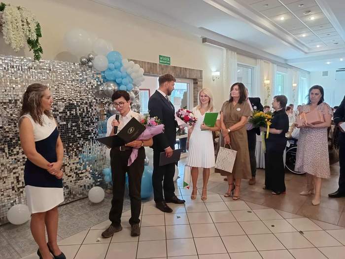 Pokaż zdjęcie: Grupa kilku osób stoi w kolejce do składania życzeń oraz wręczania kwiatów i upominków. Jedna z kobiet trzyma w ręce mikrofon i odczytuje list gratulacyjny skierowany do innej kobiety, która stoi na tle ściany udekorowanej balonami i kwiatami.