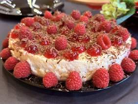 Pokaż zdjęcie: Apetyczny deser w formie sernika, udekorowany świeżymi malinami, wiśniami kandyzowanymi oraz startą czekoladą. Deser podany jest na czarnym talerzu, a w tle widać ozdobne detale.