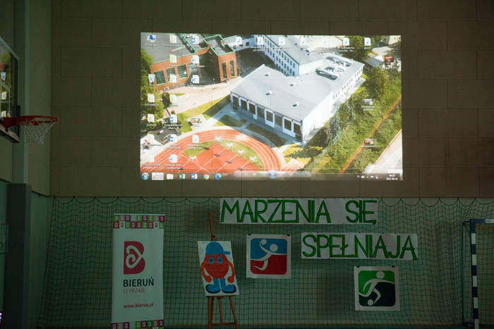 Pokaż zdjęcie: Na ścianie wyświetlany jest film pokazujący budowę nowego skrzydła budynku pod nim jest napis marzenia się spełniają i logo szkoły