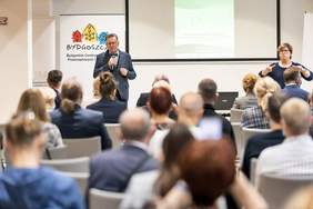 Pokaż zdjęcie: na sali siedzą uczestnicy forum - tyłem do kamery, na drugim planie widać Wicemarszłaka województwa Zbigniewa Ostrowskiego, który przemawia, obok tłumaczka języka migowegho, w tle baner Bydgoskiego Centrum Organizacji Pozarządowych i Wolontariatu 