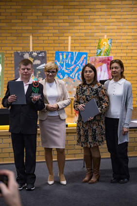 Pokaż zdjęcie: Cztery osoby stoją obok siebie na tle wystawy kolorowych prac plastycznych. Mężczyzna po lewej stronie trzyma dyplom i statuetkę w kształcie tulipana. Obok niego stoją trzy kobiety – dwie z dokumentami w rękach. Wszyscy pozują do zdjęcia podczas gali konkursowej.