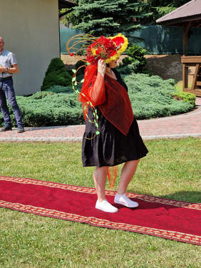 kobieta w czarnej sukience i sporym kapeluszu czerwono-żółtym i czerwonej chuście idzie po czerwonym dywanie