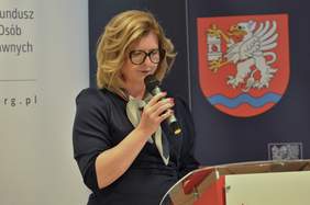 Pokaż zdjęcie: Przy mównicy stoi kobieta w ciemnych mocnych okularach.