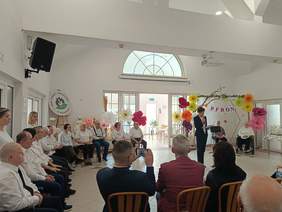 Pokaż zdjęcie: Zdjęcie z uroczystości w jasnej, wysokiej sali. Uczestnicy siedzą w półokręgu wzdłuż ścian; po lewej stronie grupa mężczyzn w białych koszulach. Po prawej stronie kobieta w ciemnym garniturze przemawia do mikrofonu. Stoi obok dekoracji z wielkich, kolorowych papierowych kwiatów i gałęzi, na której widnieje napis „PFRON”. Na lewej ścianie znajduje się logo Domu Pomocy Społecznej w Baszkowie oraz czarny głośnik. Na pierwszym planie widać siedzących gości. W tle duże okna i przeszklone drzwi, przez które wpada naturalne światło.