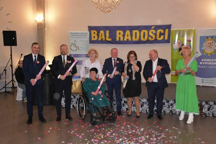 Na środku sali stoi grupa osób i trzymają w rękach tuby z konfetti, na środku, przed nimi siedzi osoba na wózku