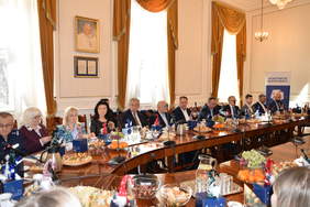 Pokaż zdjęcie: Duża sala konferencyjna. Przy owalnym stole siedzą uczestnicy posiedzenia. Kobiety i mężczyźni są elegancko ubrani. Na stol mają dokumenty, mikrofony i poczęstunek. W tle widać okna i rollup z logo OHP