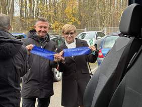 Pokaż zdjęcie: Dwie osoby stoją obok siebie, trzymając niebieską wstęgę z napisem „Powiat Białostocki – z sercem do ludzi”. W tle widać zaparkowane samochody. Widać, że wstęga została właśnie przecięta w ramach symbolicznego przekazania pojazdu.