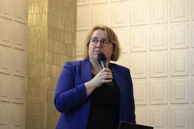 Pokaż zdjęcie: Kobieta w okularach spogląda na prezentację za nią, w ręku trzyma mikrofon. Ubrana jest w elegancką niebieską marynarkę