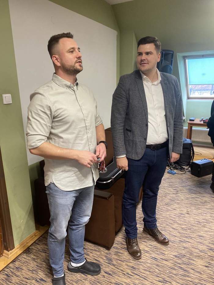 Pokaż zdjęcie: Dwóch młodych mężczyzn stoi obok siebie i rozmawia, za nimi sprzęt muzyczny