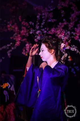 Pokaż zdjęcie: Pionowe ujęcie oświetlone fioletowym światłem, przedstawiające aktorkę w stroju stylizowanym na tradycyjne japońskie kimono. Kobieta ma białą charakteryzację twarzy (makijaż gejszy/maiko), czerwone usta i upięte włosy ozdobione kwiatem. Ubrana jest w intensywnie fioletowe kimono z szerokimi rękawami. Stoi z profilem do widza, z głową skierowaną w lewo, z dłonią uniesioną nad czołem w geście przypominającym taniec. W tle widoczne są sztuczne, różowe i białe kwiaty wiśni, tworzące ramę dla postaci.