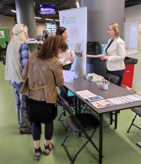 Pokaż zdjęcie: Stoisko PFRON - kobieta przy stoliku udziela informacji zainteresowanym uczestnikom targów pracy. Obok stolika stoi rollup PFRON z napisem "Wszyscy jesteśmy tacy sami" 