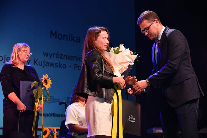 Na scenie teatru stoi Wojewoda Kujawsko-Pomorski Michał Sztybel i wręcza kwiaty i upominek  kobiecie, którą wyróżnił. W tle, po lewej stronie stoi kobieta ze słonecznikiem i dyplomem w dłoni.