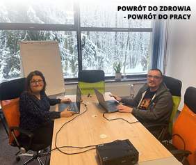 Pokaż zdjęcie: W pokoju przy dużym biurku w kolorze jasnego drewna, siedzą dwie osoby naprzeciwko siebie. Po lewej stronie siedzi kobieta, po prawej mężczyzna, obydwoje są w średnim wieku.