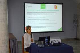 Pokaż zdjęcie: Kobieta w białej bluzce siedzi przy stole konferencyjnym i przemawia. W tle jest ekran z wyświetloną prezentacją.