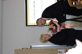 Pokaż zdjęcie: Mężczyzna w garniturze wręczający kobiecie bukiet kwiatów.)